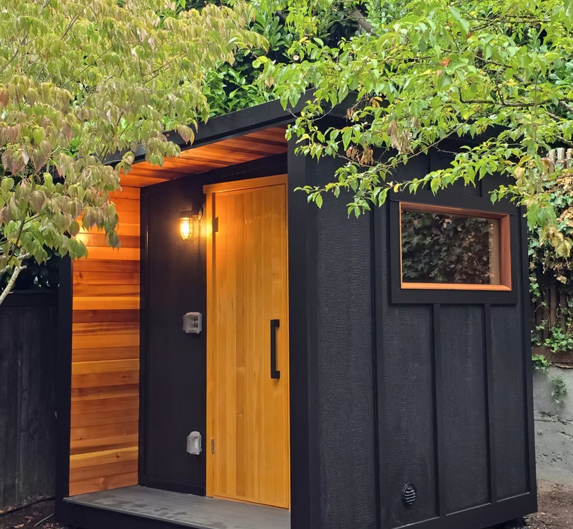 Backyard sauna clad in black board with a lit cedar door, surrounded by trees at dusk.