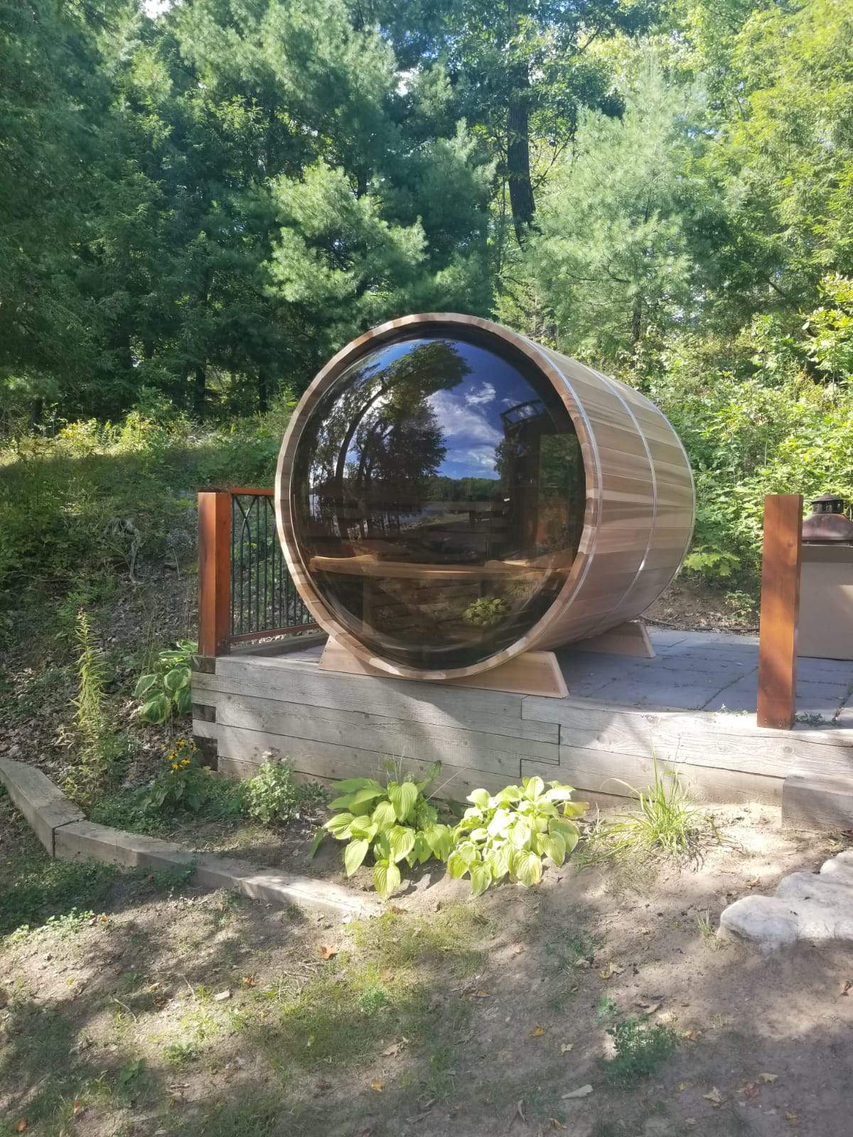 Cedar barrel sauna with a round glass end wall, set on a stone patio in a garden.