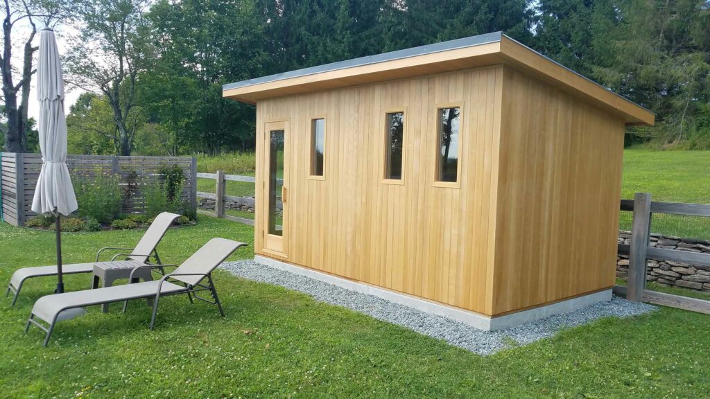 Modern flat-roofed cedar cabin sauna on a gravel pad with two loungers on the lawn beside it.