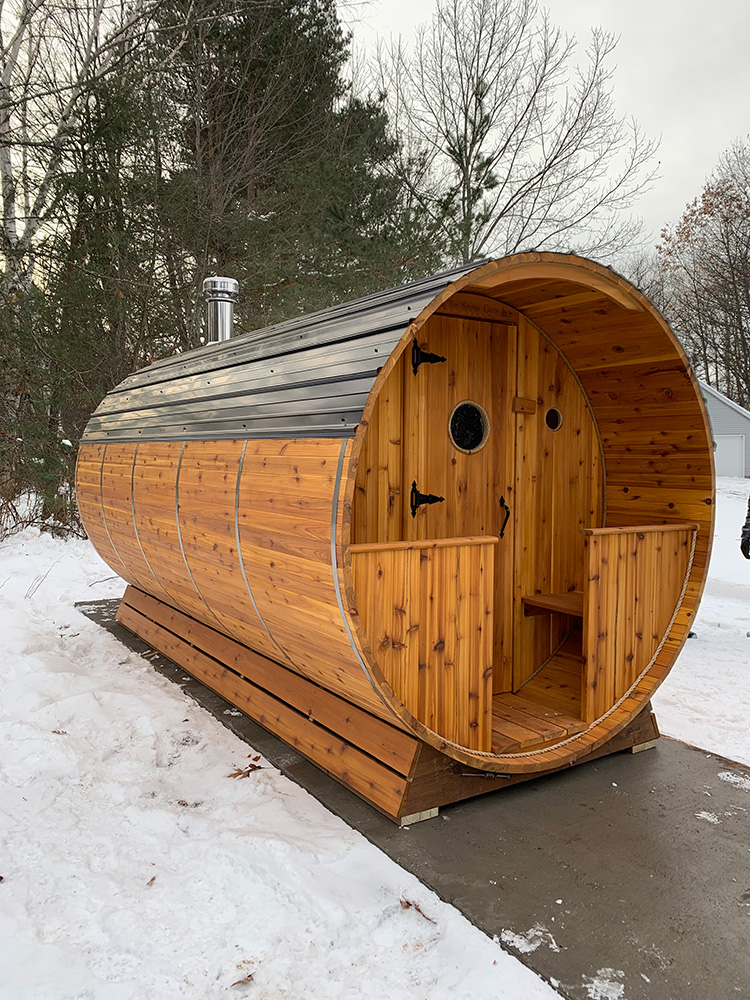 Cedar barrel sauna with a chimney standing in deep snow.